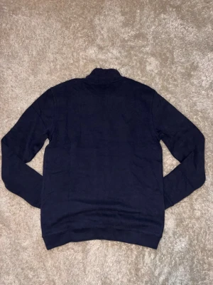 Marinblå half zip tröja Polo Ralph Lauren - Snygg marinblå långärmad tröja från Polo Ralph Lauren med half zip och ståkrage. Tröjan har ett rött broderat Polo-logo på bröstet och är tillverkad i mjuk bomull. Perfekt för dig som gillar klassisk stil med en modern touch.