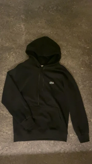 Svart Lacoste Hoodie - Säljer en svart Lacoste Hoodie med snören och luva, i nytt skick, priset kan diskuteras, frågor? Bara att kontakta!