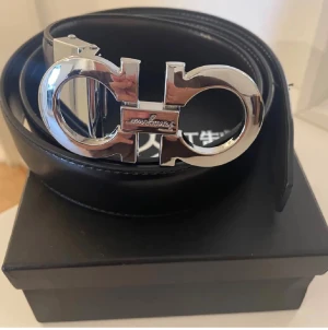 Svart skärp från Salvatore Ferragamo - Snyggt svart bälte från Salvatore Ferragamo med en ikonisk, stor silverspänne i metall. Bältet är gjort i slätt skinn och har en stilren, klassisk design som passar till många outfits. Perfekt accessoar för att lyfta din look.