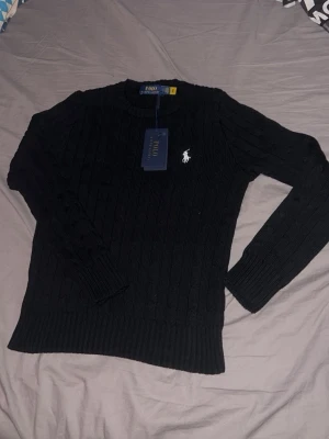 Svart stickad tröja från Polo Ralph Lauren - Kabelstickad svart tröja från Polo Ralph Lauren med rund halsringning och klassisk vit broderad logga på bröstet. Tröjan har lång ärm och ribbade muddar vid ärmslut och nederkant. Perfekt för lager-på-lager under kyliga dagar.