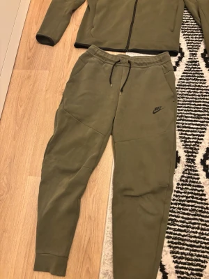 Nike tech byxa - Snygga olivgröna mjukisbyxor från Nike med svart logga på benet. Byxorna har snörning i midjan och ribbade muddar vid bensluten. Perfekta för chill eller träning, med skön passform och mjukt material.