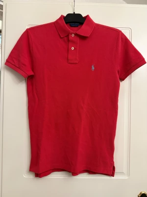 Röd piké från Ralph Lauren - Säljer en klassisk röd pikétröja från Ralph Lauren med korta ärmar och krage. Tröjan har två knappar framtill och den ikoniska blå broderade loggan på bröstet. Materialet är mjuk bomull och passformen är normal. Perfekt för en chill och stilren look.