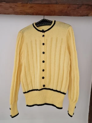 Yellow knitted sweater 70's - Yellow handmade knitted sweater in 70s style 💛 Buttons down to the waist and then a waistband. Black details in the knitting and black buttons. Soft in the yarn, and not itchy at all. Length: 65 cm, Arm length: 64 cm (from seam). I'm sure that someone can style this better than me 🫶☺️ 