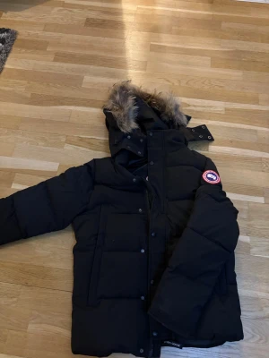 Svart Canada Goose dunjacka med päls - Svart pufferjacka från Canada Goose med ikonisk patch på ärmen, stor huva med fluffig päls och praktiska fickor. Jackan har både dragkedja och knappar framtill, perfekt för kalla vinterdagar. Jackan är använd av min lillasyster i ungefär 4 månader men den är exakt som ny.
