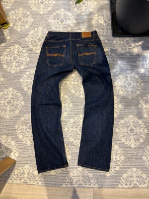Mörkblå jeans från Nudie Jeans, W30 L32 - Säljer ett par mörkblå jeans från Nudie Jeans med klassisk femficksdesign och snygga kontrastsömmar. Jeansen har raka ben och läderpatch med logga bak. Tillverkade i slitstarkt bomullsjeansmaterial. Perfekt för dig som gillar stilrena och tidlösa jeans.