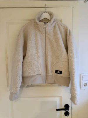 Beige teddyjacka från Adidas - Mysig beige teddyjacka från Adidas med hög krage och hel dragkedja framtill. Jackan har två stora fickor och en svart Adidas-logga på vänster sida. Perfekt för kyliga dagar och har en avslappnad passform.