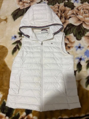 Vit dunväst från Tommy Hilfiger från Boozt - Snygg vit dunväst från Tommy Hilfiger med avtagbar huva och dragkedja framtill. Västen har två fickor med dragkedja och diskret logga på bröstet. Klassisk quiltad design och sportig känsla, perfekt för lager-på-lager-stil.