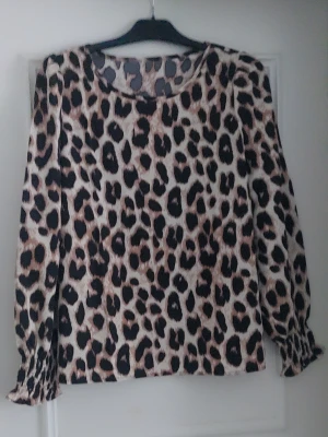 Leopardmönstrad blus med långa ärmar - Snygg blus med leopardmönster i beige, svart och brunt. Blusen har rund halsringning och långa ärmar med lätt rynkade muddar. Perfekt för dig som vill sticka ut med ett trendigt djurmönster. Materialet känns mjukt och följsamt.