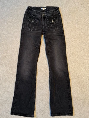 Mörkgrå bootcut Gina Tricot Young jeans med fickdetaljer - Snygga mörkgrå Gina Tricot Young jeans med bootcut passform och dekorativa fickor framtill med lock och knapp. Jeansen har klassisk femficksdesign och är tillverkade i ett mjukt denimtyg. 