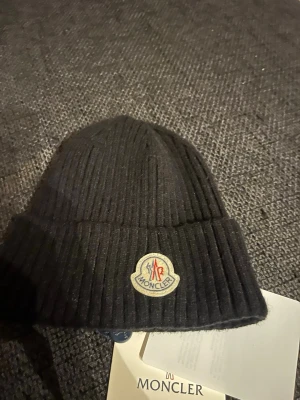 Svart ribbstickad mössa Moncler - Snygg svart ribbstickad mössa från Moncler med klassisk uppvikt kant och broderad logotyp framtill. Tillverkad i mjukt material som håller dig varm och ger en clean look. Perfekt accessoar för kalla dagar och streetstyle.