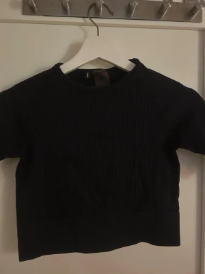 Svart ribbad crop top från Stronger - Snygg svart ribbad crop top från Stronger med korta ärmar och rund hals. Tillverkad i ett stretchigt material som passar perfekt till träning eller chill. Enkel och stilren design som är lätt att matcha med andra sportkläder.