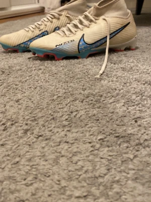 Nike Air Zoom vita fotbollsskor - Säljer ett par vita Nike Air Zoom fotbollsskor med blå detaljer och svart swoosh. Skorna har snörning, låg profil och platt sula med röda och blå inslag undertill. Perfekta för dig som vill sticka ut på planen!