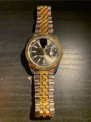 Tvåfärgad klocka från Olevs - Stilren klocka från Olevs med svart urtavla, guldfärgade detaljer och datumvisning. Armbandet är i tvåfärgad metall, guld och silver, med klassisk länkdesign. Klockan har markerade index och en räfflad guldfärgad ring runt urtavlan. Perfekt accessoar för en trendig look.