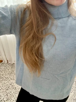 Ljusblå polotröja i mjuk stickning - Säljer en ljusblå stickad polotröja med avslappnad passform och hög krage. Tröjan är i mjukt material och har långa ärmar. Perfekt för lager på lager och enkel att matcha med jeans eller kjol.