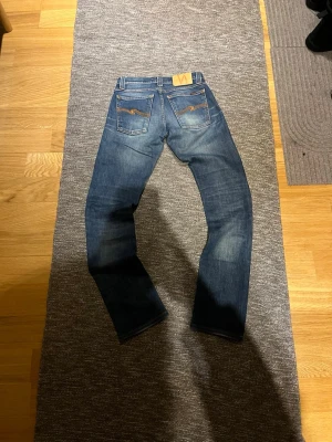 Blå jeans från Nudie Jeans Co 26/30 - Säljer ett par klassiska blå jeans från Nudie Jeans Co med snygga slitningar och kontrastsömmar. Jeansen har raka ben, fem fickor och Nudie-logga på bakfickan. Strl 26 i midjan benlängd 30. Skicka prisförslag 