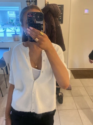 Vit kortärmad kofta med pärlknappar - Snygg vit kofta med korta ärmar och pärlknappar framtill. Koftan har en mjuk och fluffig känsla och en avslappnad passform. Perfekt att slänga över ett linne för en stilren look.