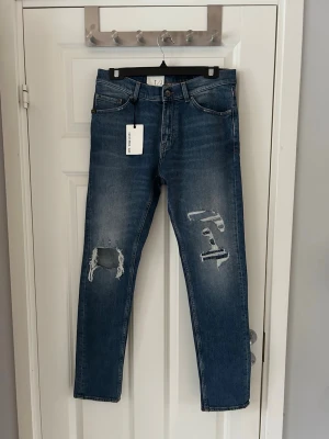 Blå slitna jeans från tiger of sweden - Snygga blå jeans från tiger of sweden med slim fit passform och slitna detaljer på båda benen. Jeansen har klassisk femficksdesign, knappgylf och är tillverkade i denim. Perfekta för en avslappnad och trendig look.