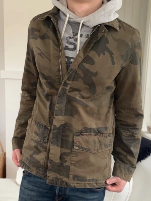 Camouflage vindjacka i bomull - Snygg vindjacka med klassiskt camouflage-mönster i gröna och bruna toner. Jackan har två stora fickor framtill, krage och stängs med dragkedja. Tillverkad i bomull och har en avslappnad passform, perfekt för en cool streetstil.