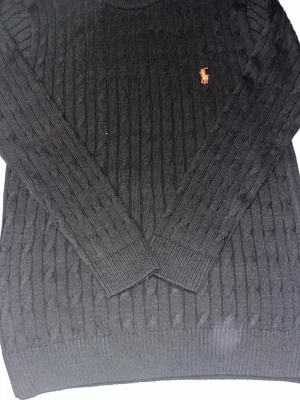 Svart kabelstickad tröja Polo Ralph Lauren - Kabelstickad svart tröja från Polo Ralph Lauren med rund halsringning och den klassiska röda logotypen broderad på bröstet. Tröjan har ribbade muddar vid ärmslut och nederkant. Perfekt för dig som gillar stilrena och tidlösa plagg.