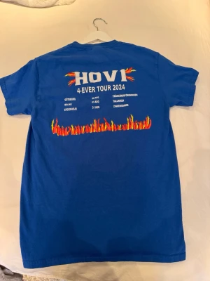 Blå Hov1 tour t-shirt 2024 - Blå t-shirt från Hov1 med tryck från 4-EVER TOUR 2024 på ryggen och en liten röd krona på bröstet. T-shirten har korta ärmar och är gjord i mjuk bomull. Snygg och enkel design med eld-detaljer och turnédatum på baksidan. OBS tröjan jag fotat är min privata tröja, tröjan jag kommer att skicka är den som ligger inplastad. Alltså aldrig öppnad eller provad!! 