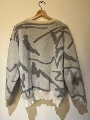 Grå stickad tröja - Stickad tröja i grömed abstrakt mönster. Tröjan har rund halsringning, långa ärmar och ribbade muddar vid ärmslut och nederkant. Perfekt för dig som gillar en avslappnad och trendig stil.