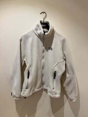 Vit fleecejacka från The North Face - Mysig vit fleecejacka från The North Face med hög krage och dragkedja framtill. Jackan har två fickor med dragkedja och en broderad logga på bröstet. Storlek M men mer som en S.