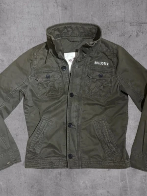 Olivgrön overshirt från Hollister - Stilren olivgrön overshirt från Hollister med fyra fickor framtill, knappstängning och hög krage. Tillverkad i bomullstyg med diskret logga på bröstet. Perfekt lager-på-lager-plagg för en avslappnad look. Den är 9/10 för dragkedjan fungerar inte men man tänker inte på det för man använder ändå knappar men annars är allt annat perfekt 