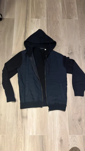 Mörkblå cardigan med dragkedja - Säljer en mörkblå cardigan med dragkedja och huva. Utsidan är quiltad på kroppen medan ärmarna och huvan är i mjuk bomull. Ribbstickade muddar vid ärmslut och nederkant. Perfekt för en chill och avslappnad stil.