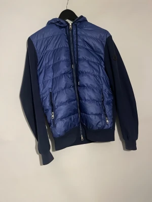 Blå quiltad pufferjacka med huva - Snygg blå hybridjacka med vadderad framsida och huva. Ärmar och rygg är i stickat material medan framsidan är quiltad och glansig. Två dragkedjefickor framtill och hel dragkedja. Perfekt för lager-på-lager och streetstyle. Xl men passar S/M