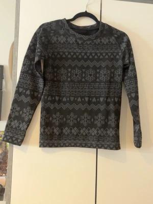 Svart tröja med snöflingemönster - Mysig svart långärmad tröja med grått snöflinge- och hjärtmönster. Tillverkad i 96% polyester och 4% elastan för en stretchig och bekväm passform. Perfekt för kalla dagar och snygg till jeans eller mjukisbyxor.