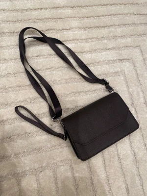 Svart axelväska från H&M - Stilren svart axelväska från H&M i klassisk och tidlös design. Perfekt storlek för mobil, plånbok och andra vardagsnödvändigheter – smidig att bära både till vardags och vid tillfällen då du vill ha en enkel men snygg väska. Väskan har justerbar axelrem, lock med tryckknapp samt innerficka med dragkedja som gör det lätt att hålla ordning. Använd men i fint begagnat skick.  Märke: H&M Färg: svart