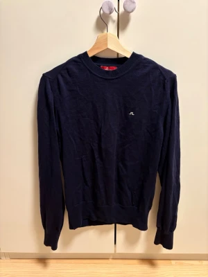 J.Lindeberg Stickad merino tröja - Snygg mörkblå stickad tröja från J.Lindeberg med diskret logga broderad på bröstet. Tröjan har rund halsringning, långa ärmar och ribbade muddar vid ärmslut och nederkant. Perfekt till lager på lager och enkel att matcha.