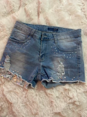 Blå jeansshorts med paljettdetaljer - Snygga blå low waist jeansshorts med slitningar, paljettdetaljer och råa kanter. Shortsen har klassisk femficksdesign och är dekorerade med små glittriga stenar framtill. Perfekta för sommaren och ger en cool, trendig look.