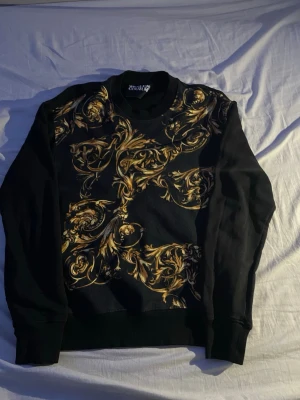 Svart sweatshirt med guldmönster - Cool svart sweatshirt med unikt barockinspirerat guldmönster på framsidan. Tröjan har rund halsringning, långa ärmar och ribbade muddar vid ärmslut och nederkant. Perfekt för dig som vill sticka ut med en trendig och stilren look.