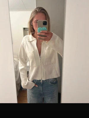 Vit ziptröja med krage och fickor - Snygg vit ziptröja med krage och två stora bröstfickor. Tröjan har en rak passform och långa ärmar, perfekt att slänga över en t-shirt. Enkel och stilren design som passar till jeans eller kjol.