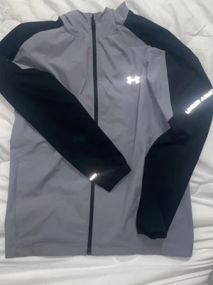 Under Armour set  - Snygg grå vindjacka från Under Armour med svarta ärmar och hel dragkedja framtill. Jackan har hög krage, reflekterande logga på bröstet och text på ärmen. Perfekt för blåsiga dagar och sportiga outfits.