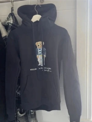 Svart hoodie Polo Bear Ralph Lauren - Svart hoodie från Ralph Lauren med den ikoniska Polo Bear-trycket på bröstet. Hoodien har huva med dragsko och ribbade muddar. Materialet ser ut att vara mjukt och bekvämt, perfekt för en avslappnad stil. Snygg detalj med björnen i preppy outfit.