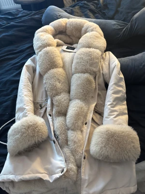 Vit pälsjacka med fluffig krage - Säljer en supermysig vit pälsjacka med stor fluffig krage och breda pälskanter vid ärmsluten. Jackan har dragkedja framtill och två fickor med dragkedja. Perfekt för kalla dagar när du vill vara både varm och snygg.