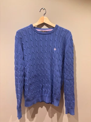 Blå kabelstickad tröja från Morris  - Snygg blå kabelstickad tröja med rund hals och ett litet broderat emblem på bröstet. Tröjan har långa ärmar och ribbade muddar vid ärmslut och nederkant. Perfekt för dig som gillar klassisk och stilren design. Tröjan är i nyskick. Storlek M.