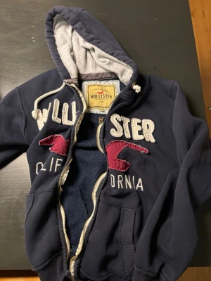 Marinblå Hollister hoodie med dragkedja - Passformen är mer som en M/S än L. Mysig marinblå hoodie från Hollister med dragkedja, broderad logga och text i vitt samt röda detaljer på bröstet. Utrustad med huva och vita dragsnören, samt två fickor framtill. Insidan är fodrad i mjukt material för extra komfort. dragkejdan är lite konstig, men den funkar ändå.