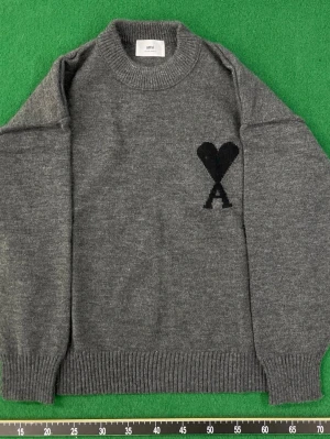 Grå stickad tröja från Ami Paris - Snygg grå stickad tröja från Ami Paris med svart hjärta och bokstaven A broderat på bröstet. Tröjan har rund hals, långa ärmar och ribbade muddar. Perfekt för dig som gillar stilrena och trendiga plagg i mjukt material.
