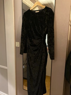 Svart sammetsklänning med rynk - Elegant svart långklänning i mjuk sammet med långa ärmar och snygg rynkning  i midjan. Klänningen har en klassisk rund halsringning och en smickrande passform som ger en lyxig känsla. Den är öppen på sidan. 