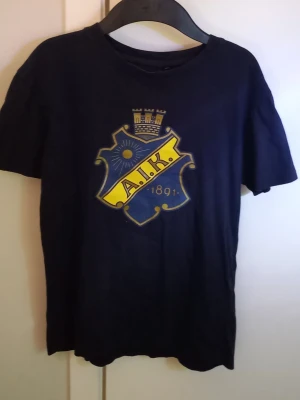 AIK t-shirt i mörkblå bomull - Snygg mörkblå AIK t-shirt med klubbens klassiska logga tryckt på bröstet i gult och blått. Tillverkad i mjuk bomull och officiell merchandise. Perfekt för dig som vill visa ditt AIK-stöd på ett stilrent sätt. Storlek 160