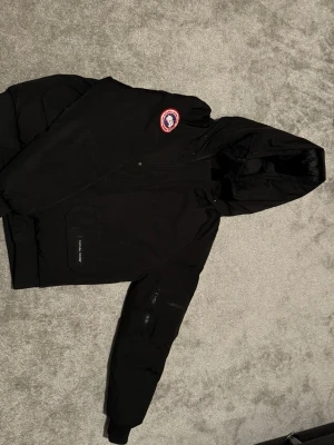 Svart dunjacka från Canada Goose - Säljer en svart dunjacka från Canada Goose med huva och klassisk logga på bröstet. Jackan har dragkedja, stora fickor och quiltad insida. Perfekt för kalla dagar och riktigt snygg streetstil.
