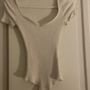 Vit ribbad body med v-ringning - Säljer en vit ribbad body med v-ringning och korta ärmar. Toppen har en tight passform och är tillverkad i ett stretchigt material som sitter snyggt på kroppen. Andvänt en gång