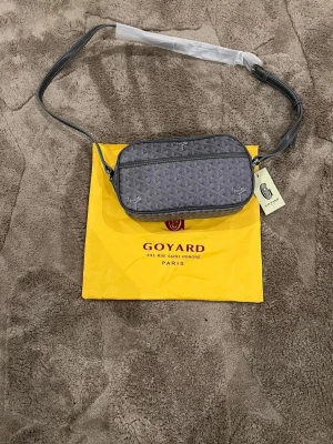 Grå axelväska  - Snygg grå axelväska med klassiskt mönster och justerbar axelrem. Väskan har en rektangulär form och detaljer i vitt och svart. Perfekt för dig som vill ha en stilren och exklusiv accessoar. Skriv innan köp och priset kan ändras om det finns möjlighet för ett direkt köp 