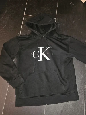 Svart hoodie från Calvin Klein Jeans - Snygg svart hoodie från Calvin Klein Jeans med stor vit CK-logga och text på bröstet. Klassisk modell med huva och dragsnören samt en stor magficka. Perfekt för en chill och stilren look. Tillverkad i mjukt bomullsmaterial. Se bild 2 för slitning på text men det är inget som stör.
