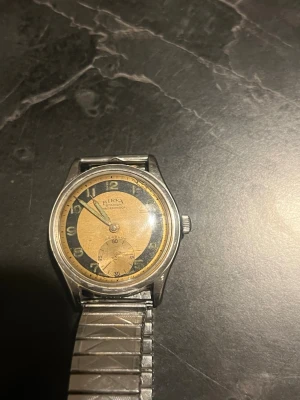 Birka armbandsur med guldfärgad urtavla - Klassisk armbandsklocka från Birka med rund guldfärgad och svart urtavla, tydliga siffror och sekundvisare. Boetten är i silverfärgad metall och armbandet är ett flexibelt metallband. Perfekt för dig som gillar retrostil och unika accessoarer.