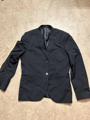 Svart kavaj från H&M Regular Fit - Klassisk svart kavaj från H&M i Regular Fit-modell. Kavajen har två knappar framtill, smala slag och fickor med lock. Perfekt för en stilren och enkel look. Tillverkad i ett slätt material med helfodrad insida.