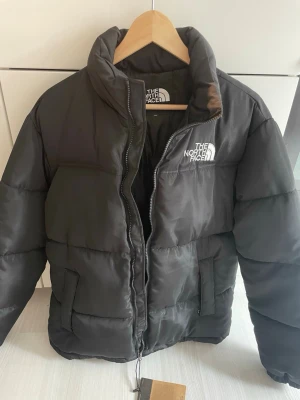 Svart pufferjacka från The North Face - Snygg svart pufferjacka från The North Face med hög krage och dragkedja framtill. Jackan har två sidofickor och logga broderad på bröstet. Perfekt för kalla dagar och riktigt trendig look. Storlek M. 